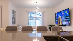 A modern living room with a large flat-screen TV mounted on the wall, a dining table with chairs, and a large window with blinds. - Amazing House Near the Parks (Kissimmee)