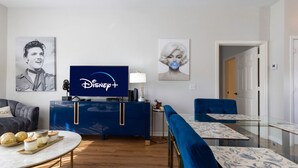Olohuoneessa on sininen viihdekeskus, jossa on litteänäyttöinen televisio, jonka näytöllä näkyy Disney+-logo, koriste-esineillä koristeltu sohvapöytä ja sininen sohva.