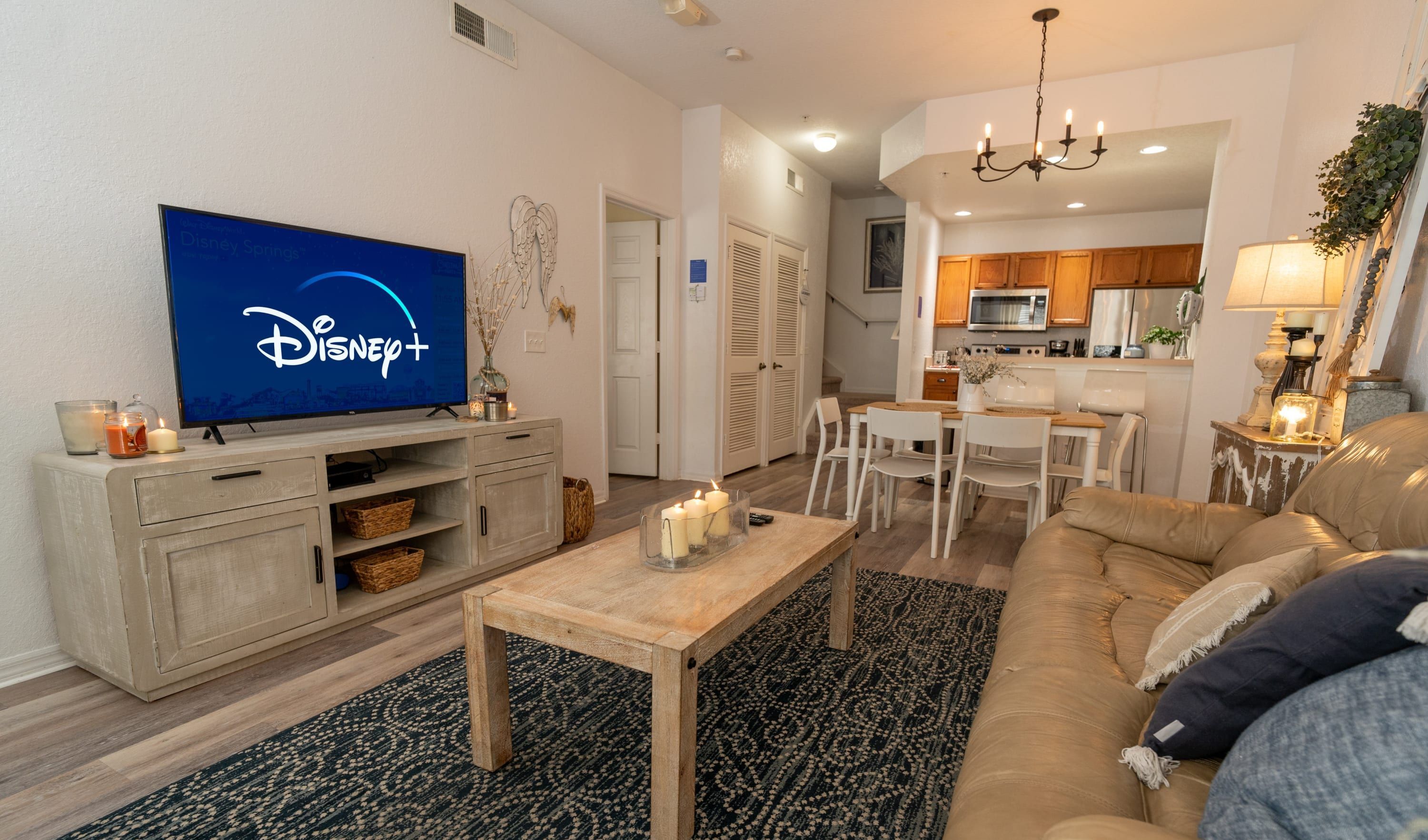 Un salon avec un téléviseur diffusant du contenu de Disney+, une table basse en bois, un tapis à motifs, un canapé beige et, au fond, une cuisine.