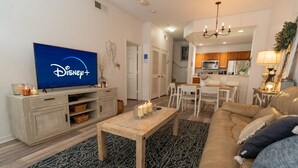 A living room with a television displaying Disney+, a wooden coffee table, a patterned rug, a beige sofa, and a kitchen in the background. - Apartment Minutes From Disney (Kissimmee)