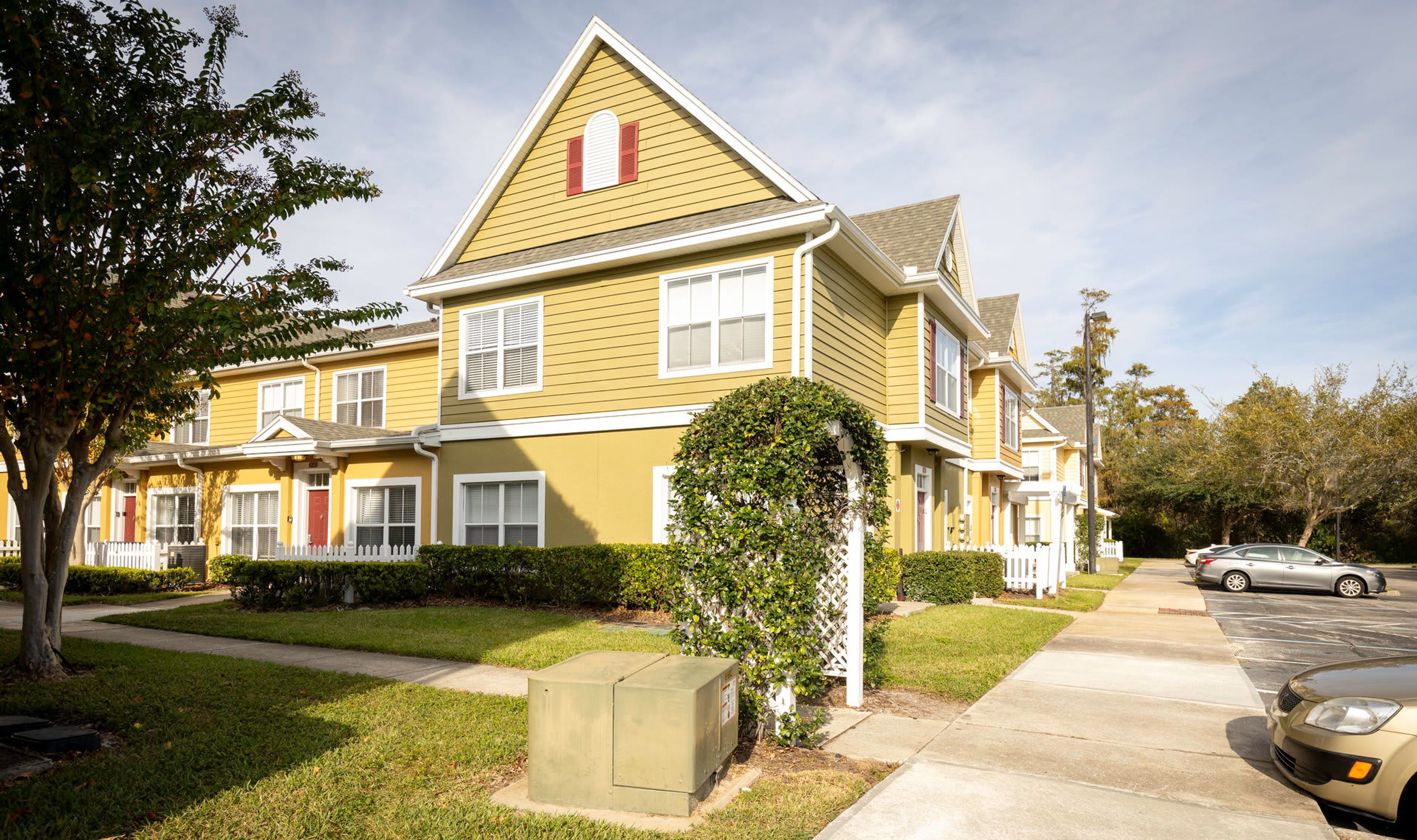 A yellow multi-story residential building with a white picket fence, a small garden, and a parked car.