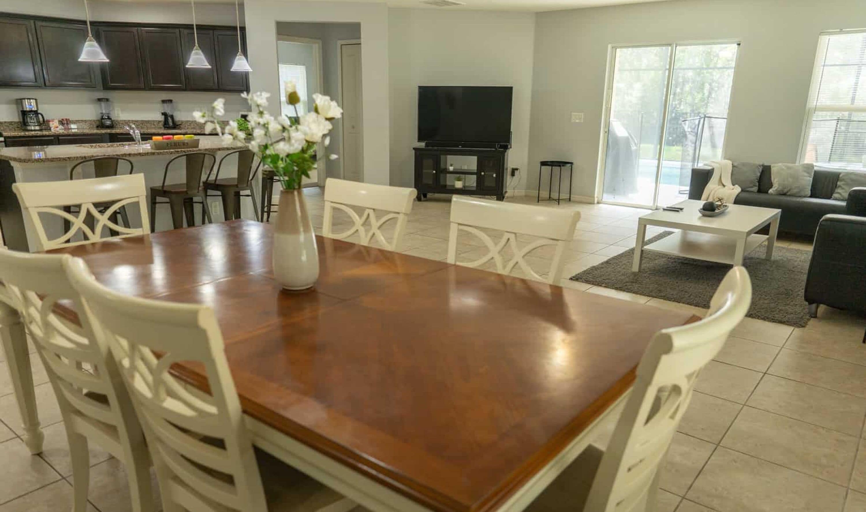 Una cocina moderna con armarios oscuros, un comedor con mesa de madera y sillas blancas, una sala de estar con sofá y televisor, y un amplio ventanal que proporciona luz natural.