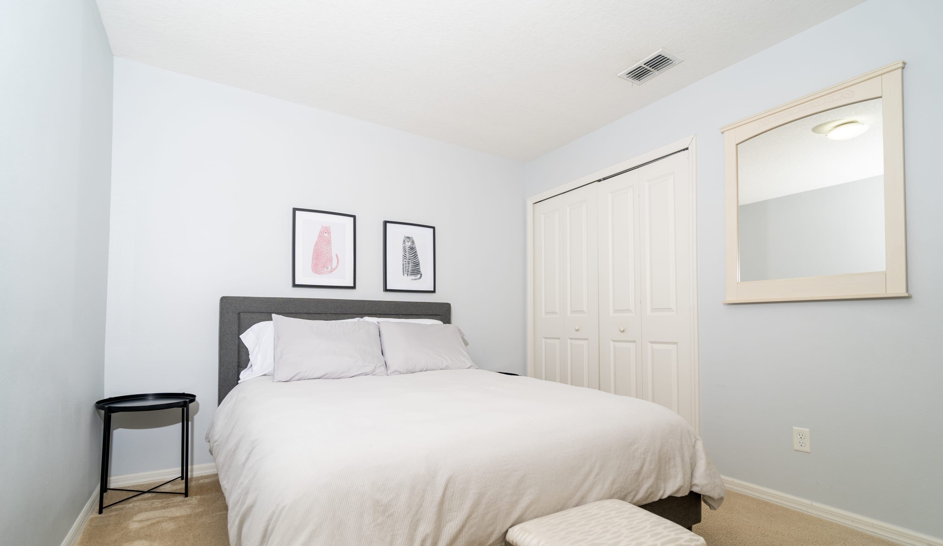 A bedroom with a bed, two framed pictures, a small table, a wardrobe, and a mirror.