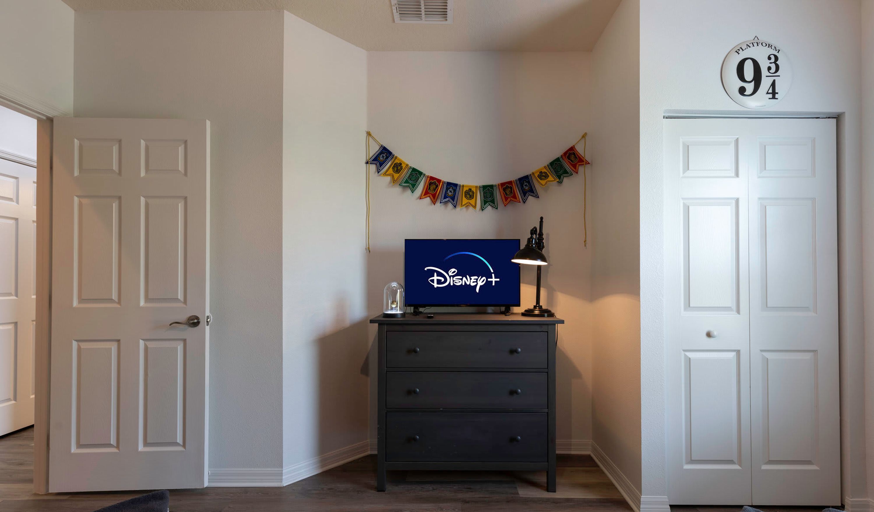 Un pasillo con un aparador de madera oscura, un televisor Disney+ encima, una lámpara y un banderín con varias banderas.