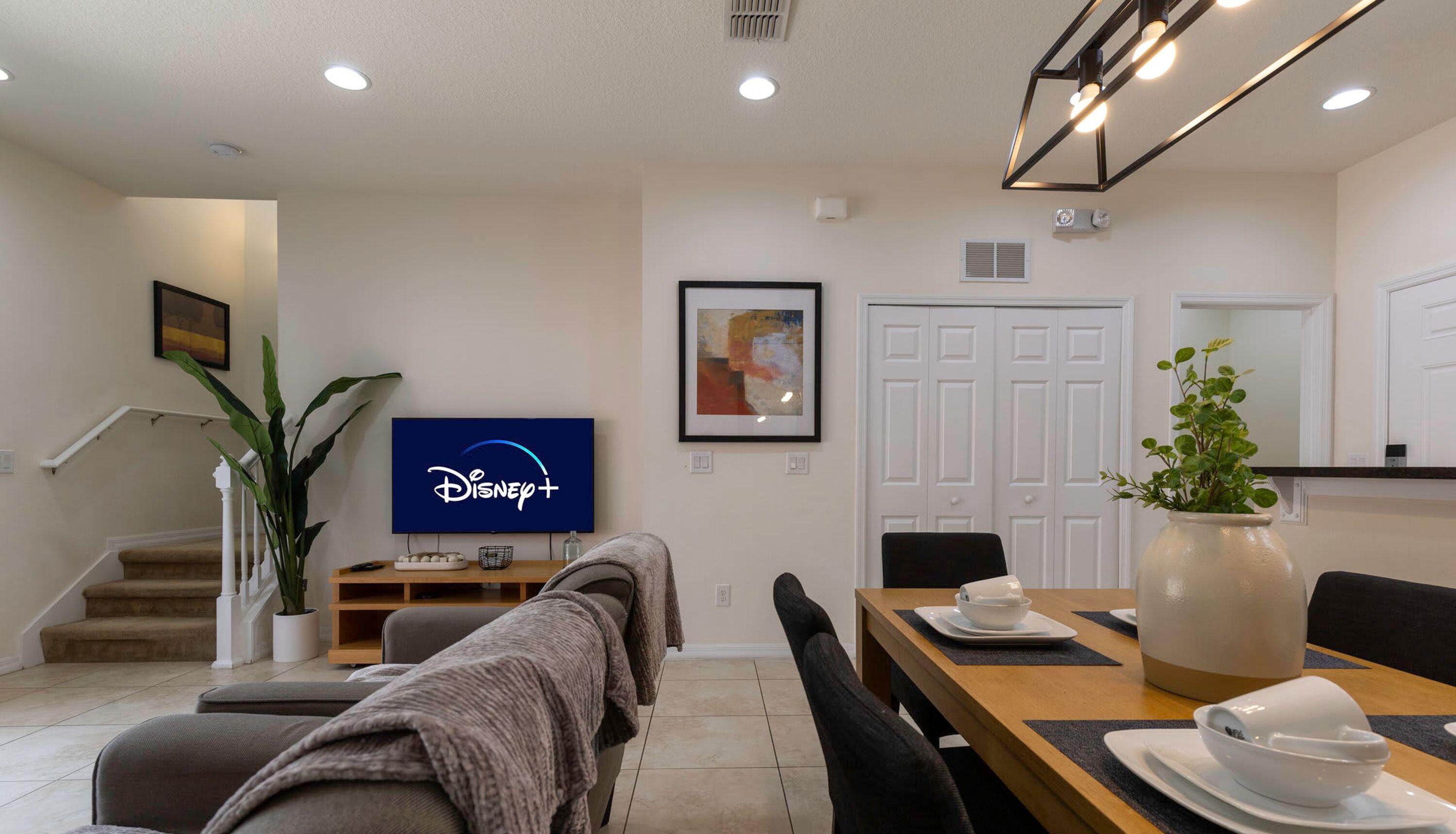 A modern living room with a flat-screen TV displaying the Disney+ logo, a dining area with a wooden table and chairs, and a staircase leading to another level.