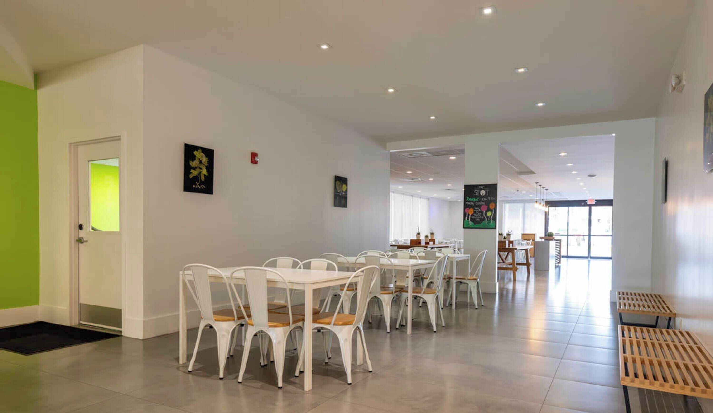 Un comedor amplio con sillas y mesas blancas, una pared verde y una puerta con ventanal.