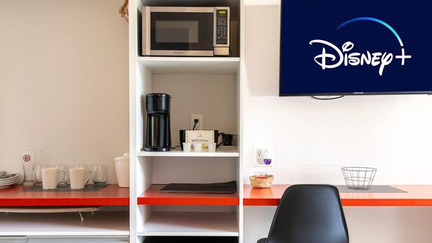 A hotel room with a kitchenette, a coffee maker, a microwave, and a television displaying the Disney+ logo. - Entire Studio With Free Shuttle to Disney (Kissimmee)