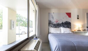 Una habitación de hotel con una cama, un cuadro abstracto grande en la pared, una ventana con vista a coches y árboles, y una mesita de noche con una lámpara.