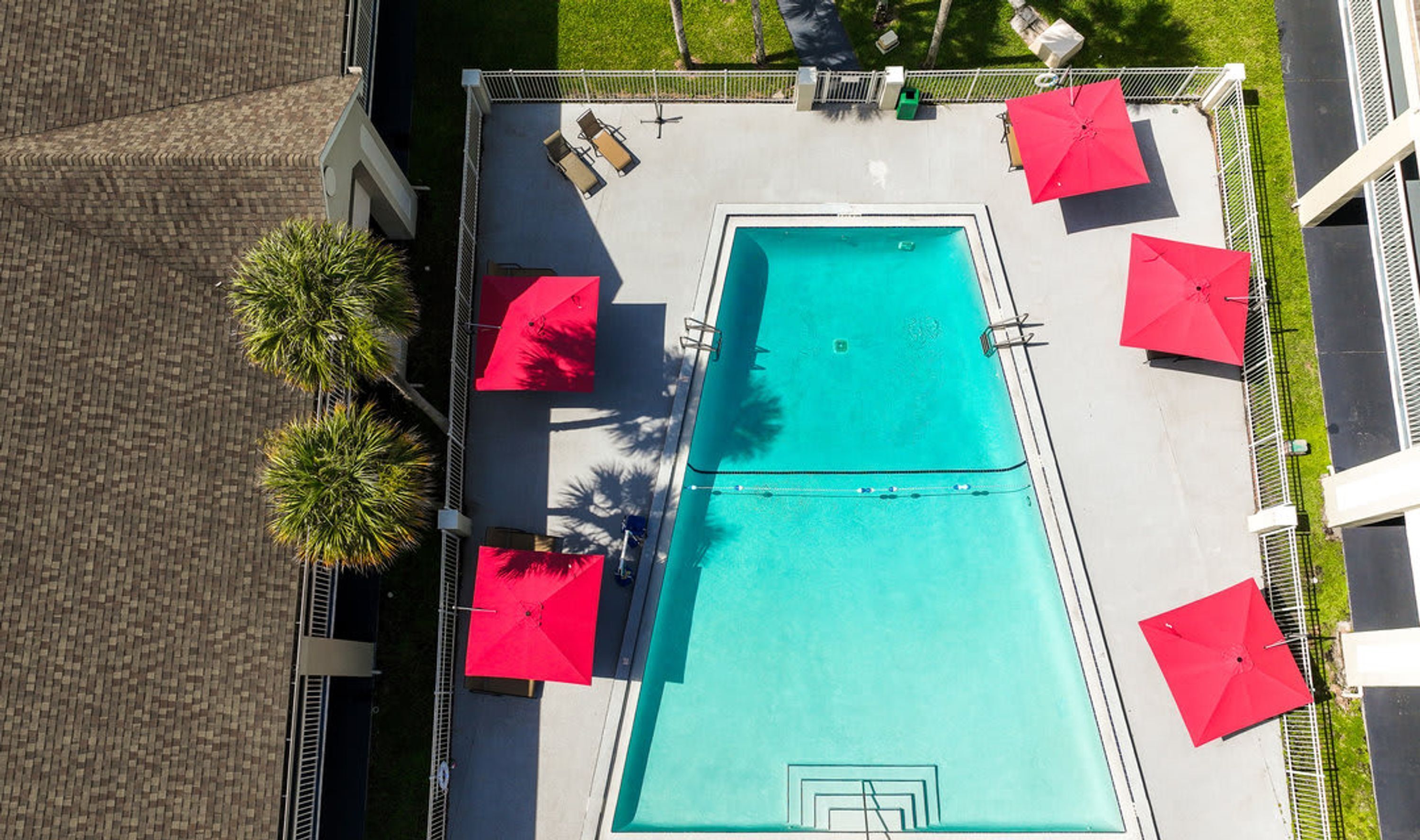 Vista aérea de una piscina rectangular rodeada por una valla blanca, con sombrillas rojas y sillas de descanso.