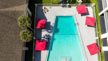 Vista aérea de una piscina rectangular rodeada por una valla blanca, con sombrillas rojas y sillas de descanso.