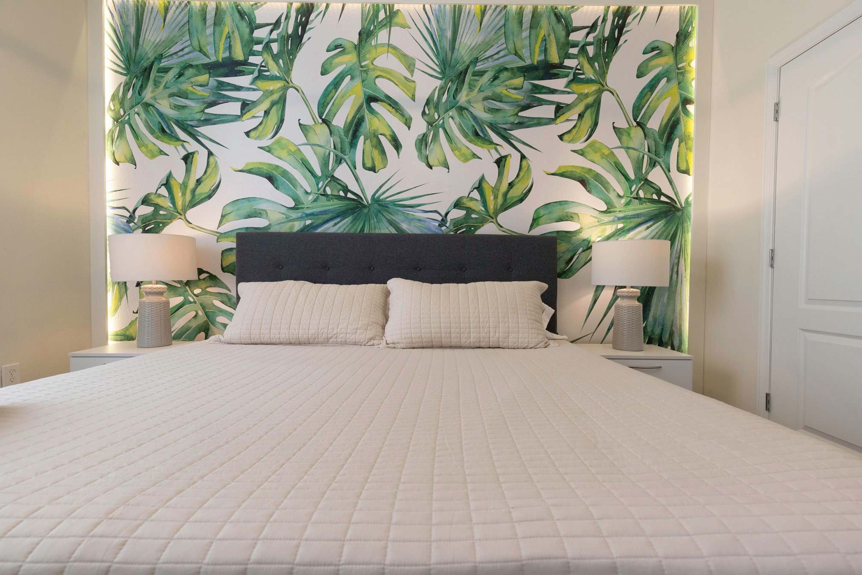 A bed with a quilted white bedspread, two white pillows, a dark headboard, and a wall with a tropical leaf pattern.