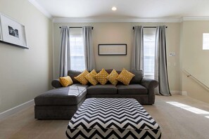 Un salon avec un canapé d’angle gris, un pouf à motifs et des rideaux gris clair.