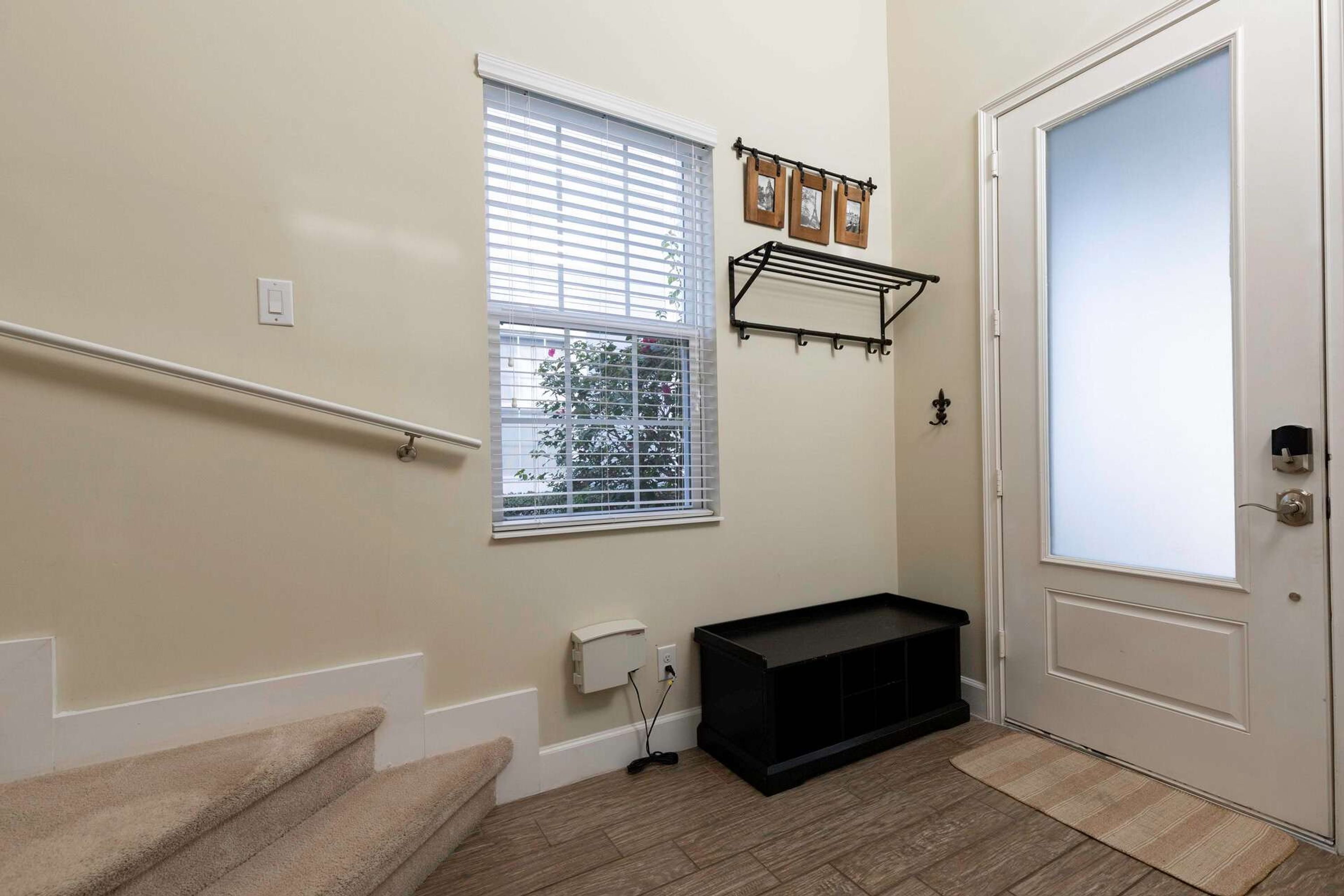 Un escalier menant à une porte, une fenêtre avec des stores, un banc noir et une étagère murale ornée de tableaux.