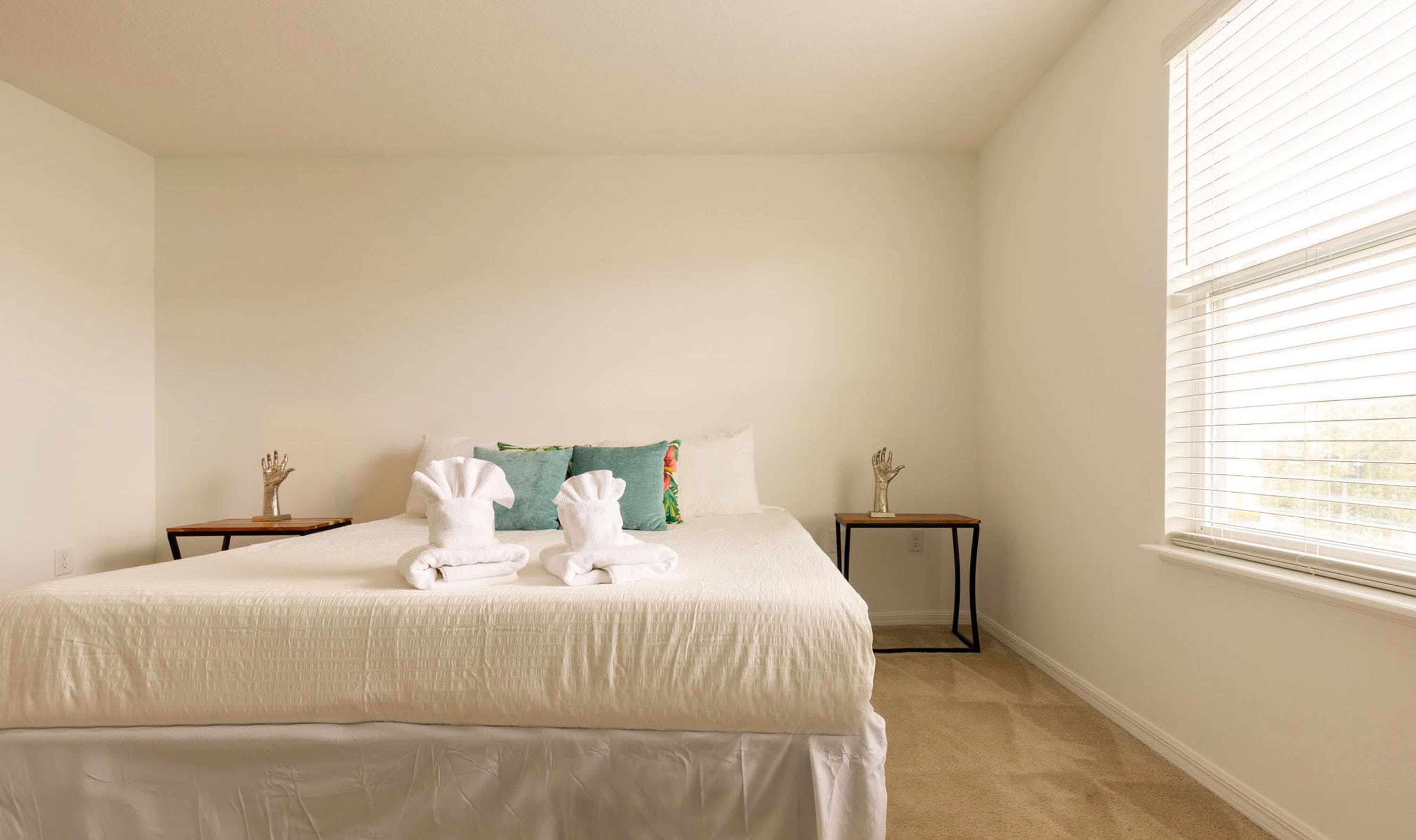 Un dormitorio con una cama grande, dos mesitas de noche, un ventanal con persianas y un cojín decorativo.