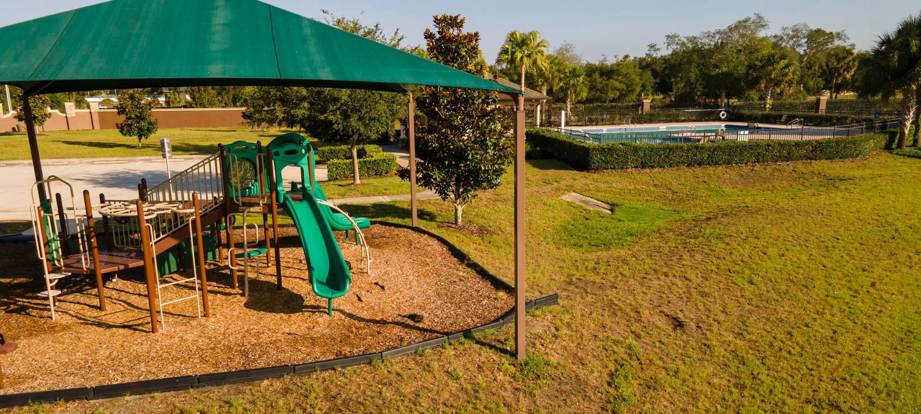 A playground with a slide and climbing structures, surrounded by a grassy area and a pool in the background.