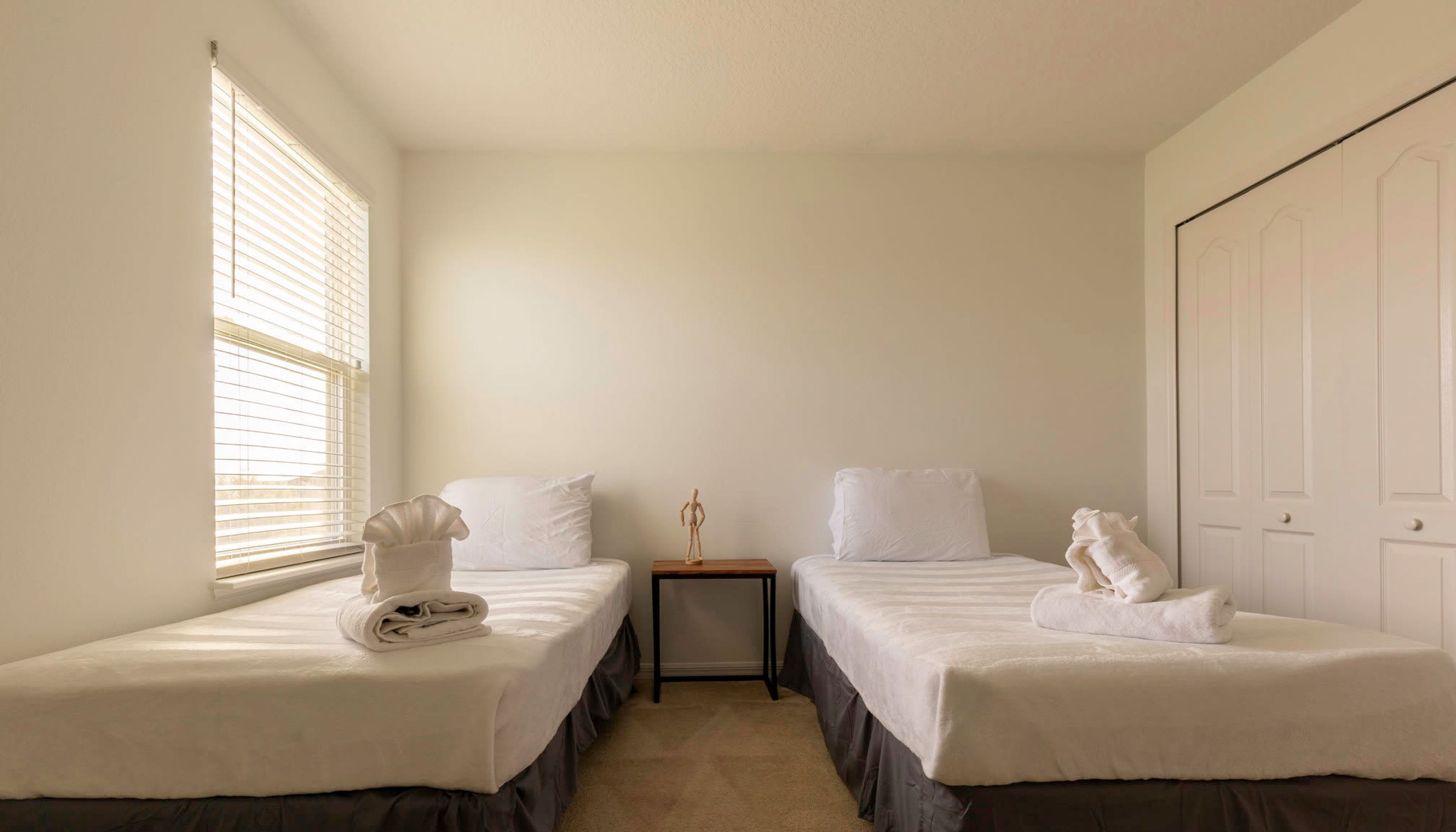 A room with two single beds, a small table, a window with blinds, and a door.