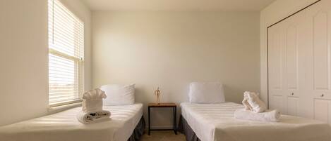 A room with two single beds, a small table, a window with blinds, and a door.