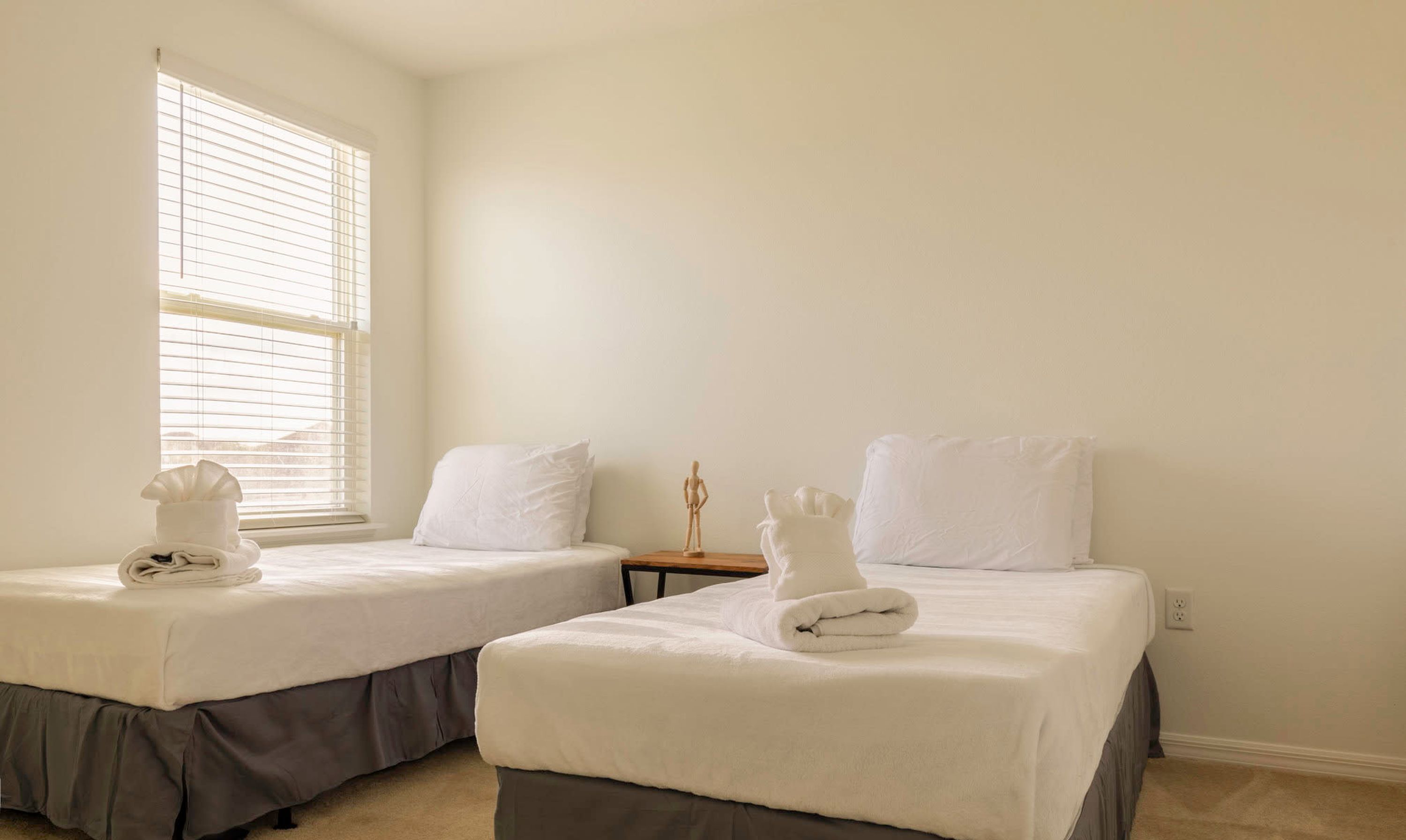 A hotel room with two beds, each with a white bedspread and pillows, and a window with blinds.