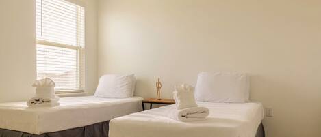 A hotel room with two beds, each with a white bedspread and pillows, and a window with blinds.