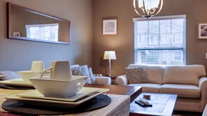 A modern living room with a sofa, a dining table with plates and bowls, a lamp, and a window with blinds. - Family Spacious 3BR Townhouse With Pool Access (Kissimmee)