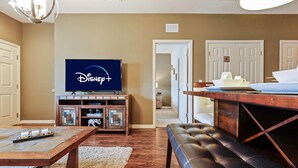 A living room with a flat-screen TV displaying the Disney+ logo, a wooden entertainment center, a dining table with a bench, and a hallway leading to a private room. - Family Spacious 3BR Townhouse With Pool Access (Kissimmee)