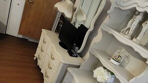 Une pièce comprenant une armoire en bois, une coiffeuse blanche avec un miroir, un téléviseur et une étagère décorative ornée d’une cage à oiseaux et de fleurs.