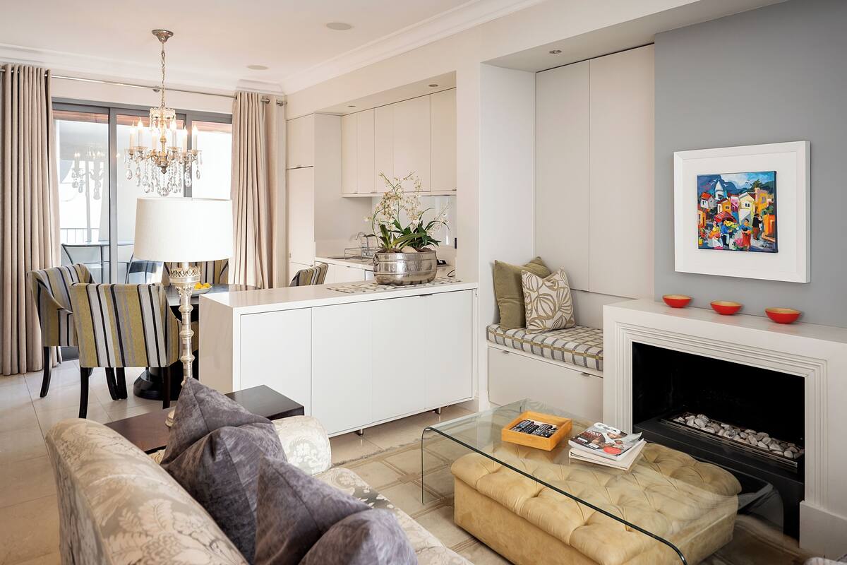 A modern living room with a sofa, ottoman, and a glass coffee table. There is a fireplace with a framed painting above it.