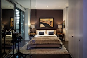 A modern bedroom with a large bed, two bedside tables with lamps, a framed artwork above the bed, and a large mirror reflecting the room. - De Waterkant Guest Suites (Cape Town)