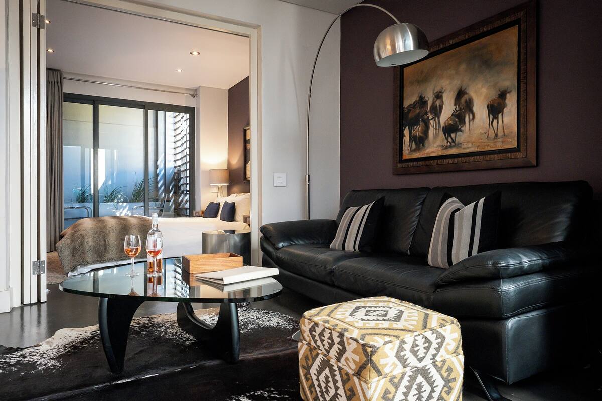A modern living room with a black leather sofa, a glass coffee table, and a patterned ottoman. A painting of deer hangs on the wall.