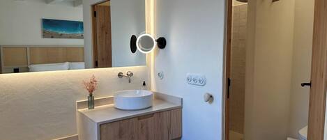 Un baño moderno con lavamanos blanco, tocador de madera y un espejo grande con luces.