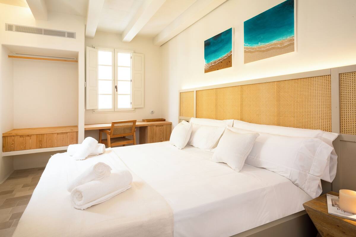 A hotel room with a large bed, a desk with a chair, a wardrobe, and two beach-themed paintings on the wall.