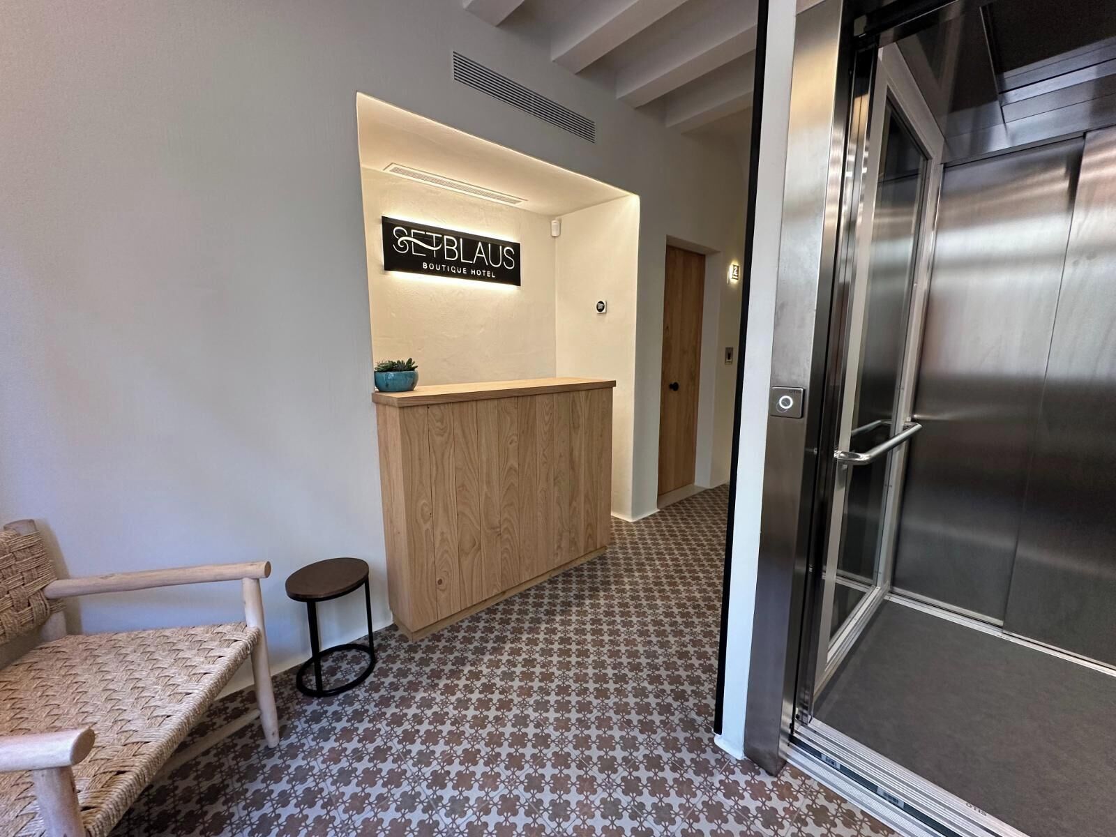 Zona de recepción de un hotel con un mostrador de madera, un pequeño rincón de descanso con un banco y un taburete, y un suelo estampado.