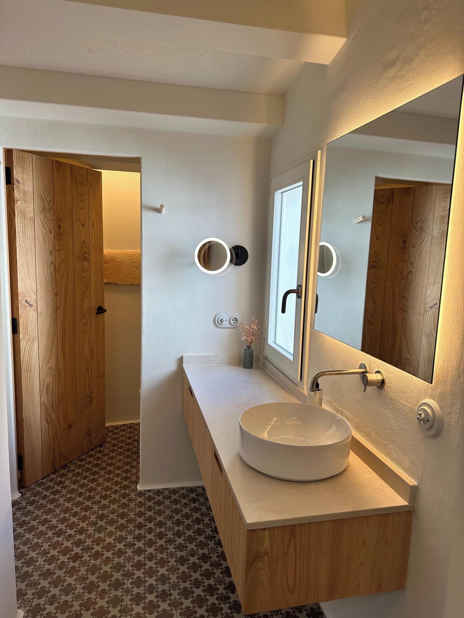 Un baño moderno con tocador de madera, un lavabo redondo, un espejo grande y un pequeño jarrón con flores.