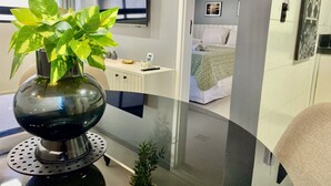 Une chambre d’hôtel moderne avec une cloison en verre, une table ronde agrémentée d’une plante en pot, un téléviseur à écran plat, une table de chevet avec un téléphone et un lit recouvert d’une couvre-lit à motifs.