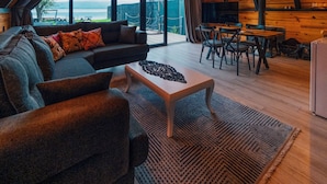 Un interior acogedor de madera con un sofá, una mesa de centro y un comedor. Amplias ventanas ofrecen vistas al exterior.