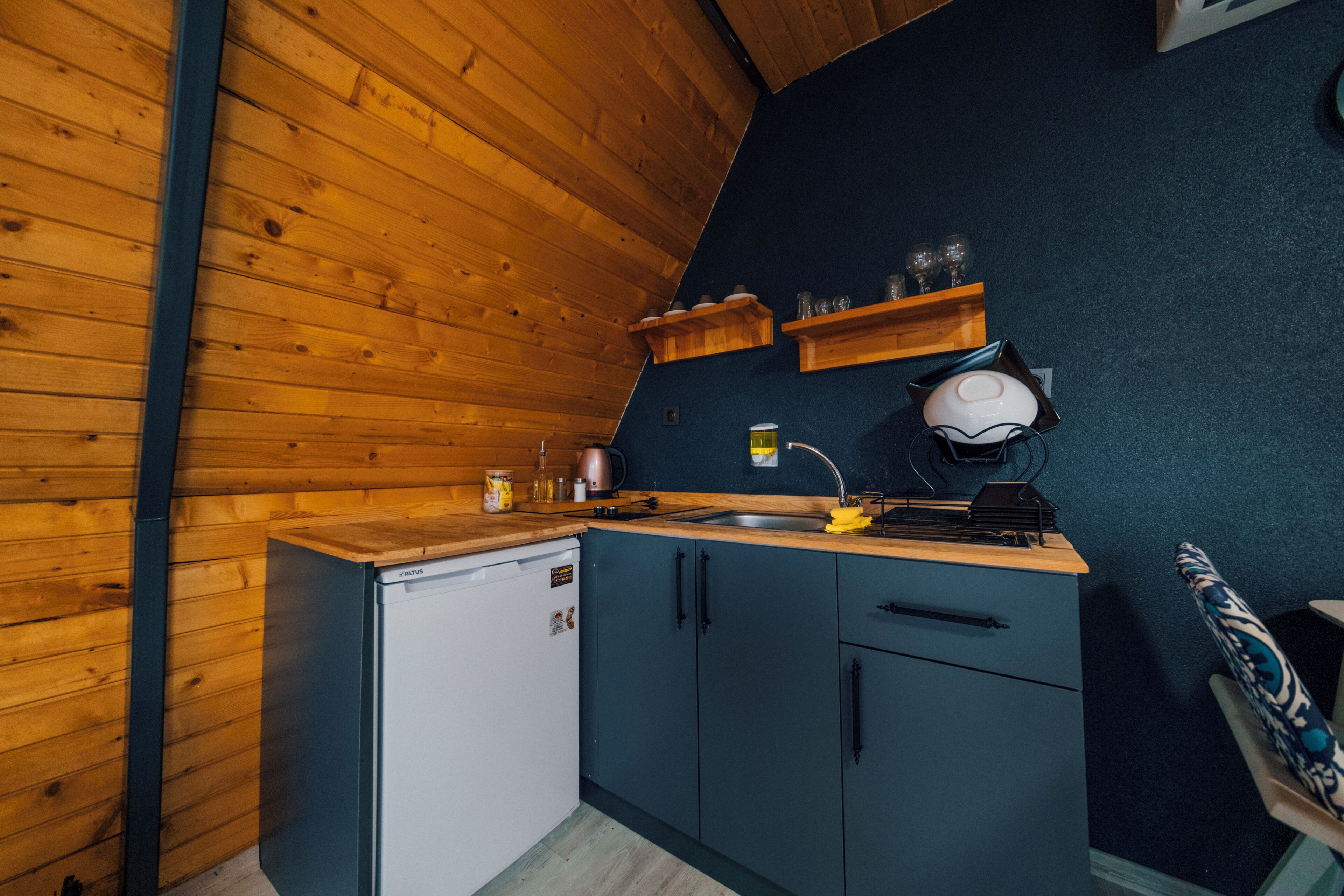 Eine kleine Küche mit Holzwänden, einem weißen Kühlschrank, dunkelblauen Schränken und einer Holzarbeitsplatte.