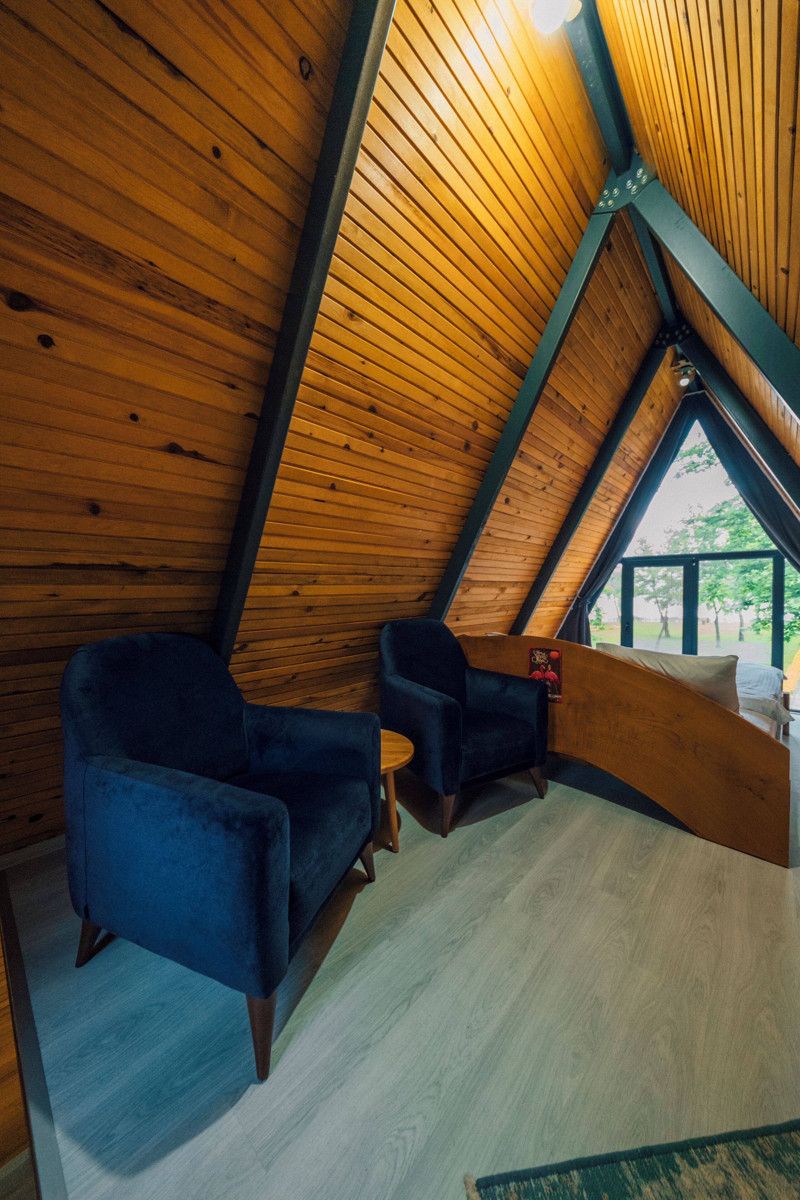 Een zolderkamer met houten latten in het plafond, twee blauwe fauteuils, een kleine tafel en uitzicht op groen.