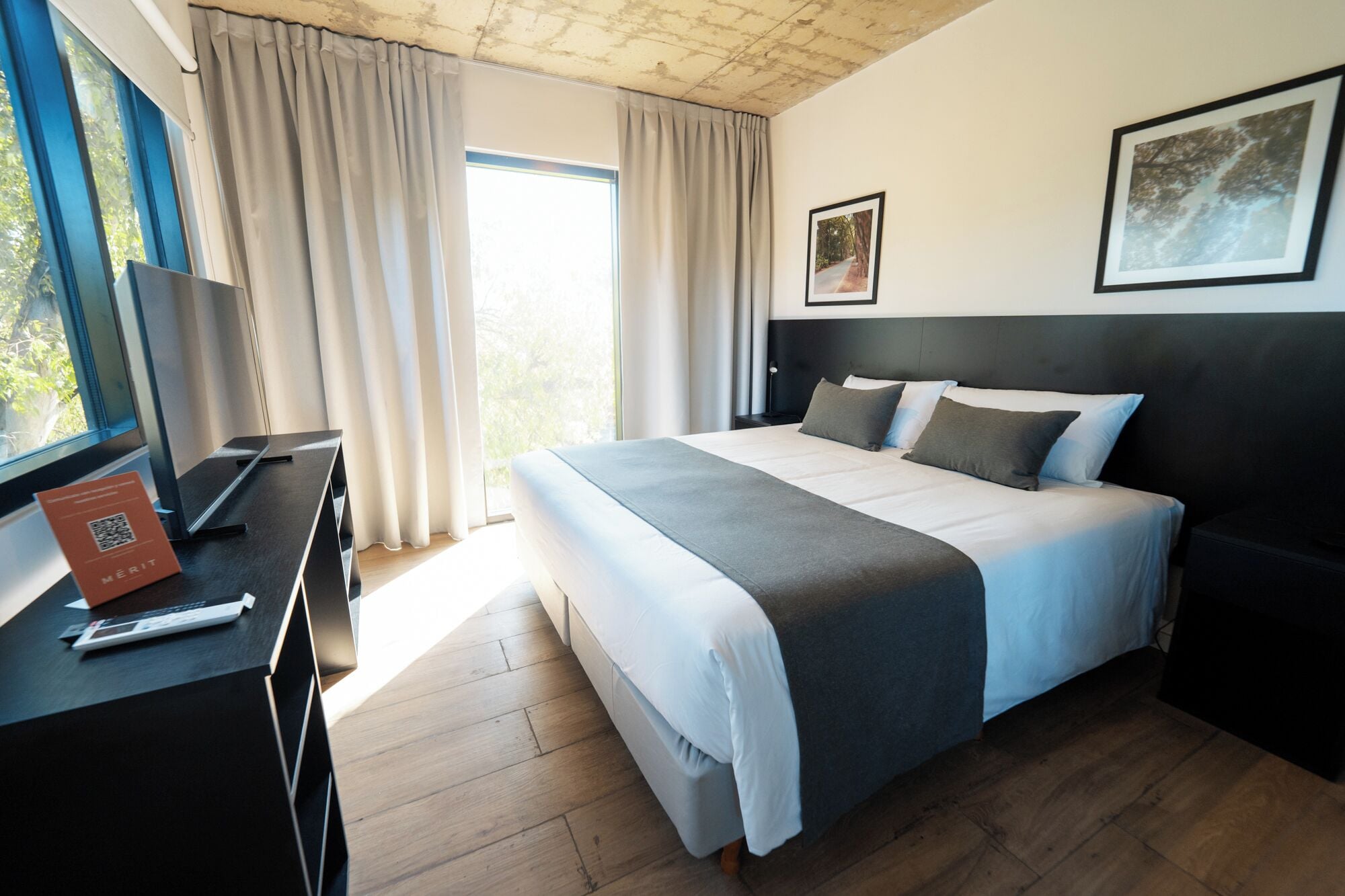 A modern hotel room with a large bed, a desk with a laptop, a window with curtains, and a framed picture on the wall.