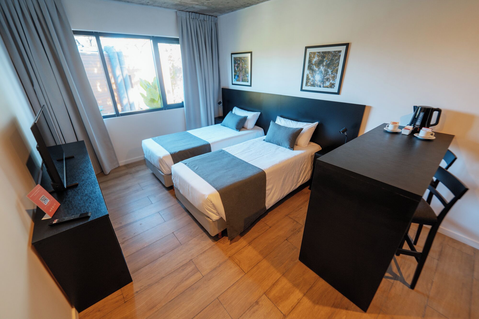 A hotel room with two beds, a desk, and a chair. There is a window with curtains, a framed picture on the wall, and a coffee machine on the desk.