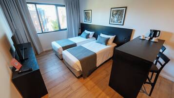 A hotel room with two beds, a desk, and a chair. There is a window with curtains, a framed picture on the wall, and a coffee machine on the desk.