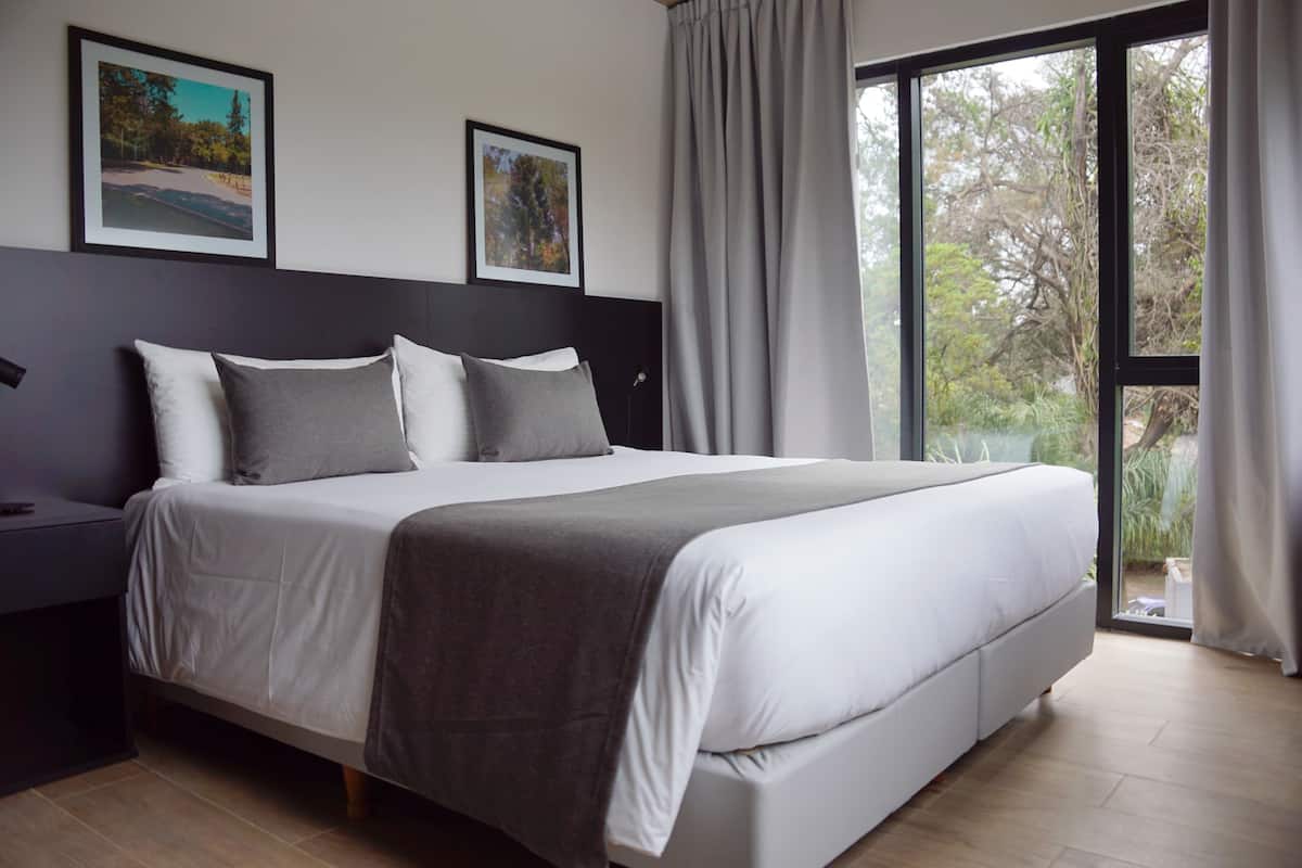 A modern bedroom with a large bed, two nightstands, a headboard, and a view of greenery through a window.