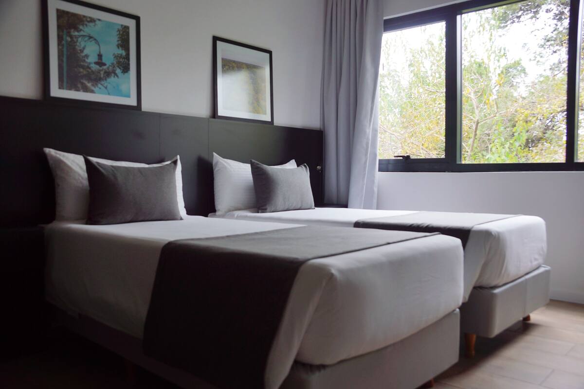A hotel room with two beds, a large window with a view of trees, and framed artwork on the wall.