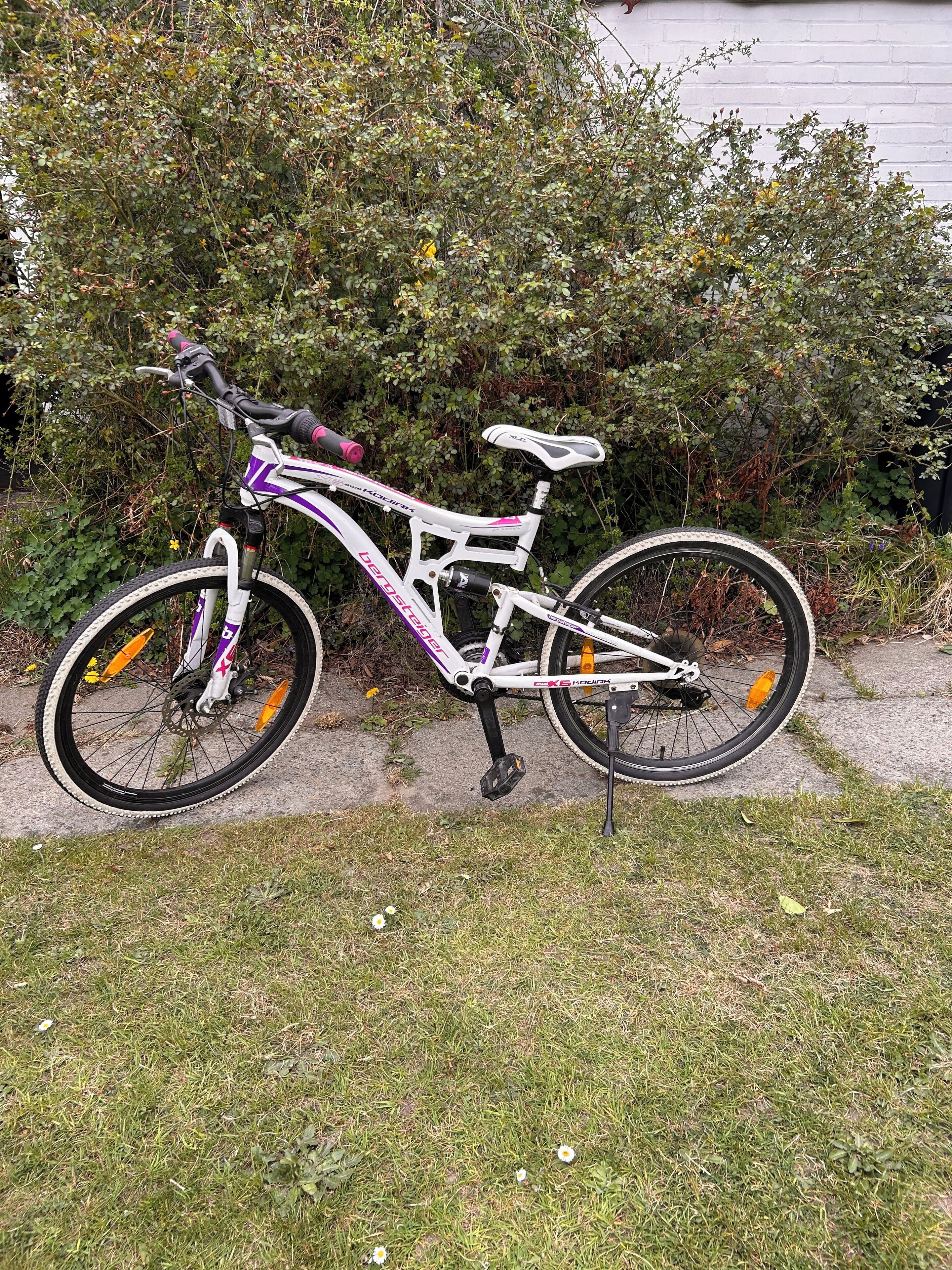 Un vélo d’appartement avec un cadre blanc et des touches de couleur violette, garé sur un chemin de béton, avec de l’herbe et un feuillage dense en arrière-plan.