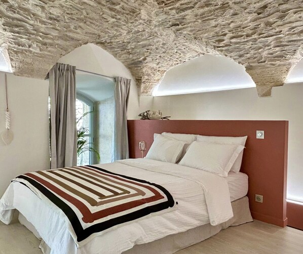 Un dormitorio con techo de piedra, una cama con ropa blanca, cabecera roja y una ventana con cortinas.
