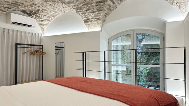 Une chambre avec un grand lit, un plaid rouge, une armoire, un miroir et une fenêtre donnant sur un paysage verdoyant.
