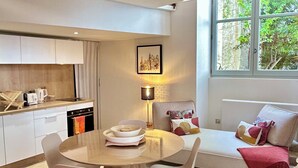 Une cuisine moderne avec des meubles blancs, une table à manger avec des chaises, un canapé avec des coussins et une fenêtre donnant sur un paysage verdoyant.