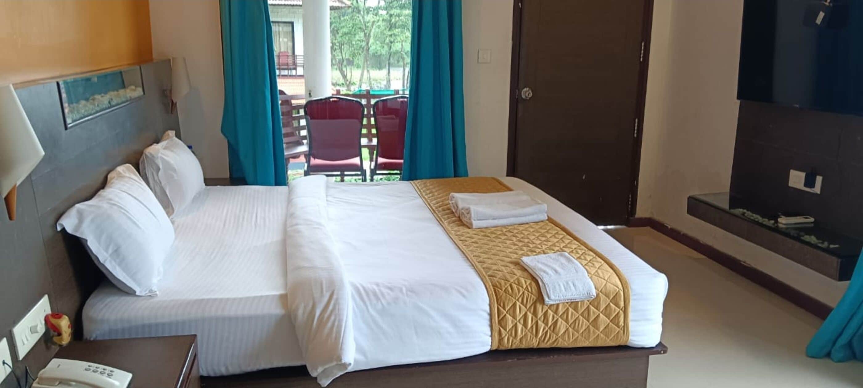 Quarto de hotel com cama, televisão, escrivaninha com telefone e vista para área externa com cadeiras e mesa.