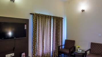A living room with a flat-screen TV, two armchairs, a coffee table with a vase, and patterned curtains.