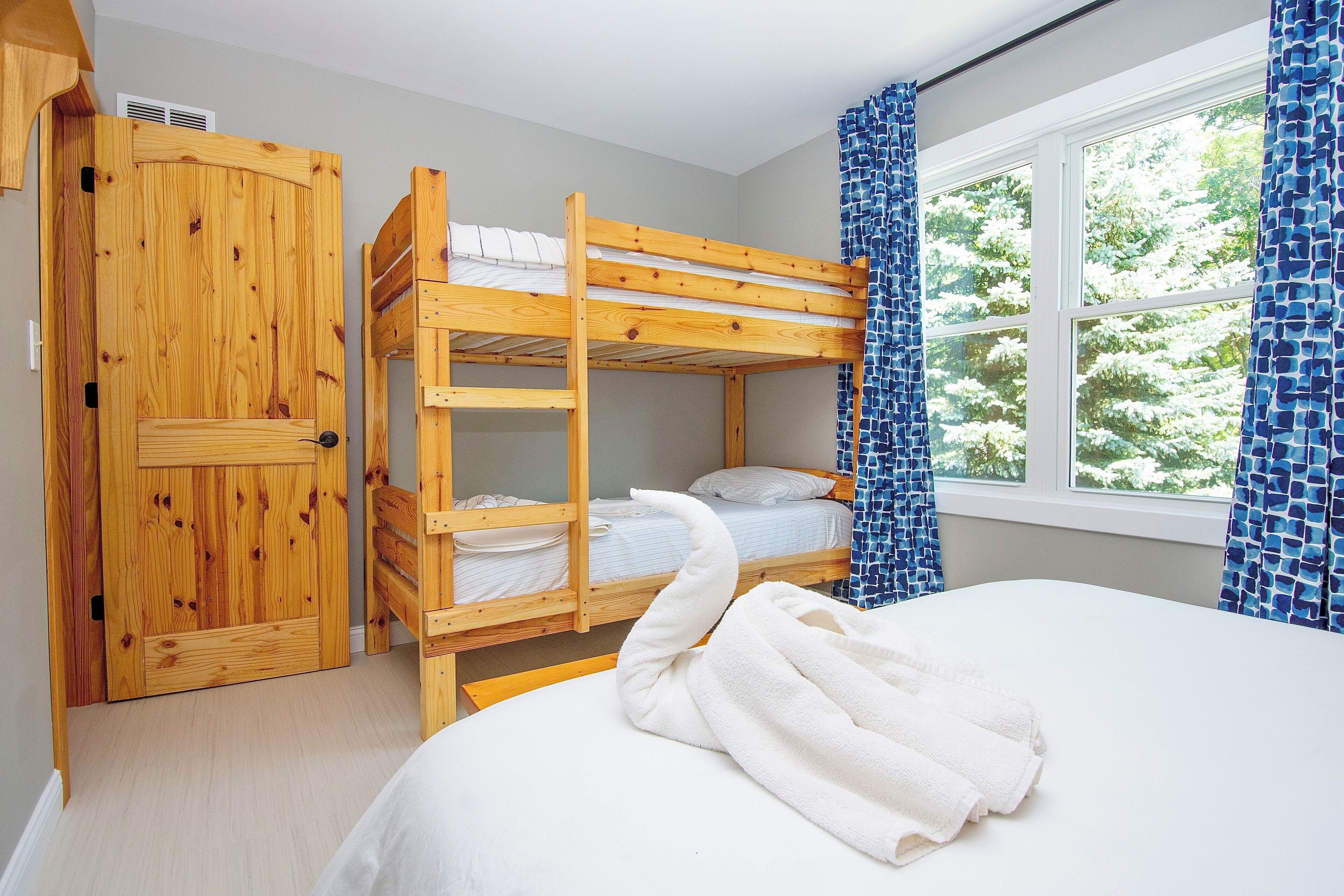 Een slaapkamer met een stapelbed, een houten kledingkast, een raam met blauwe gordijnen en een wit bed.