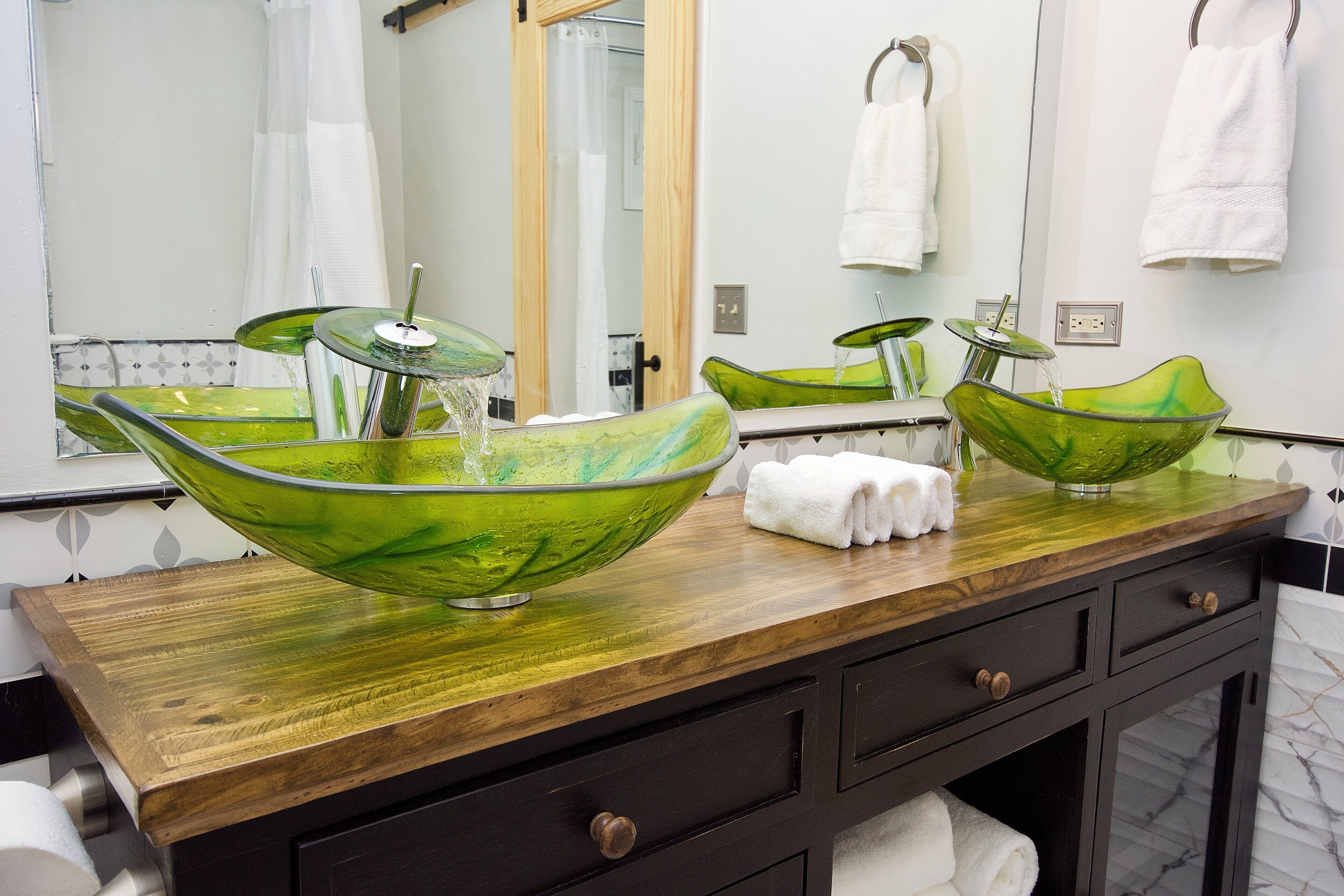 Un baño con tocador de madera, dos lavamanos de vidrio verde y un espejo.